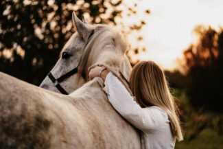 Blond young woman from behind embracing her white horse in nature in Majorca. Creative color editing with added grain. Part of a series.